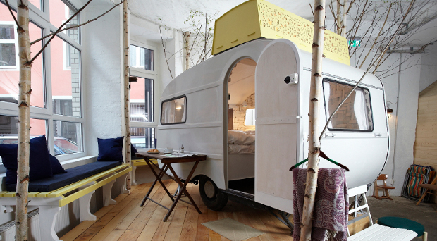 Der 70er Jahre Wohnwagen "Kleine Schwester" steht umgeben von Birken auf einem Holzpodest in der Halle.