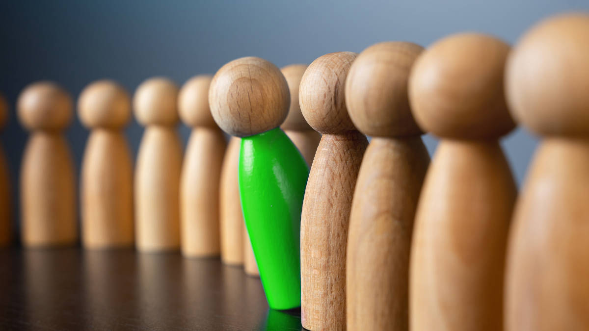 Eine Reihe identischer Holzfiguren mit einer grünen Holzfigur, die zwischen den anderen hervor schaut.