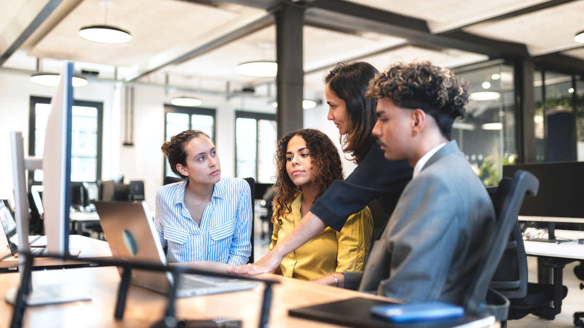 Drei junge Personen hören in einem Büro einer etwas älteren Person zu.