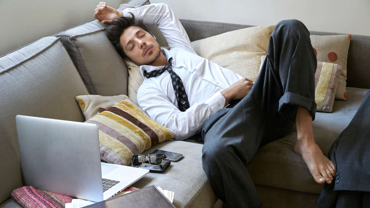 Ein Mann in Business-Kleidung schläft auf einer Couch mit seinem Notebook neben sich.