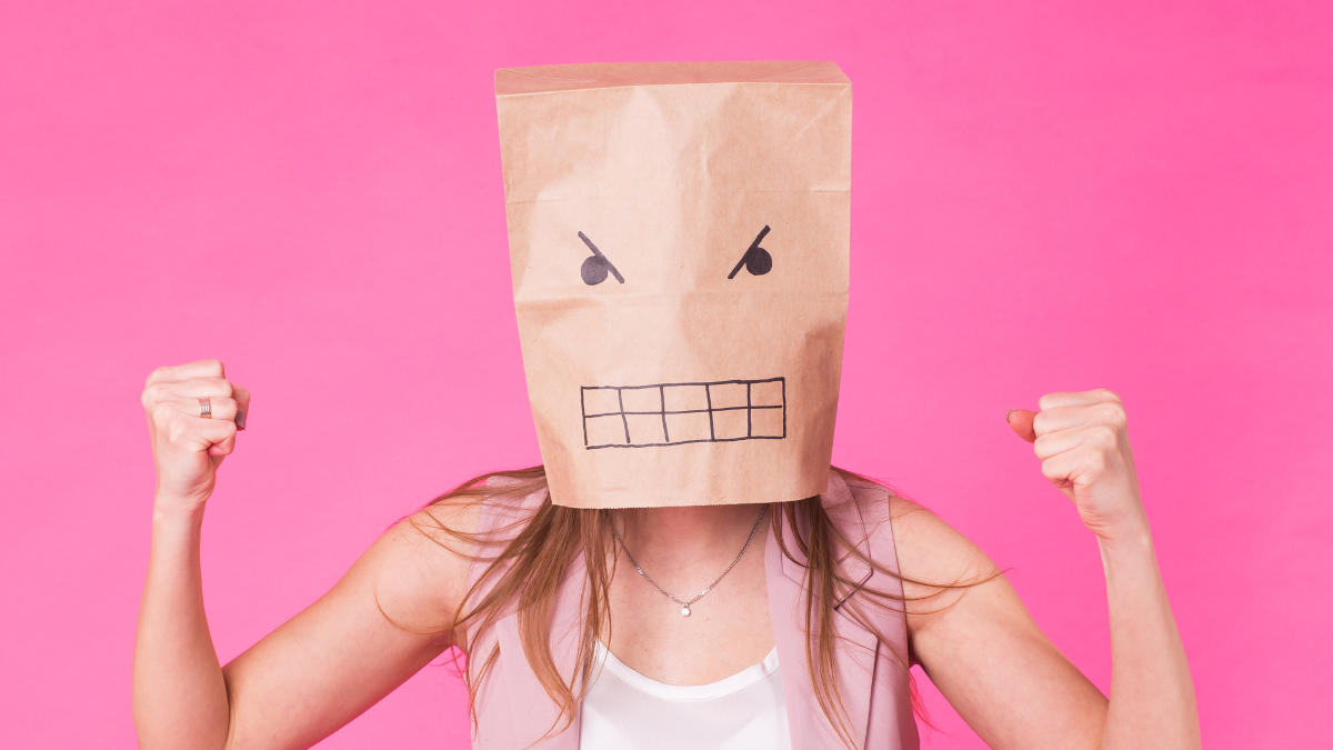 Frau mit geballten Fäusten trägt eine Papiertüte auf dem Kopf auf die ein böser Gesichtsausdruck aufgemalt ist. Sie steht vor einem rosa Hintergrund.