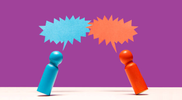 Eine rote und eine blaue Holzfigur mit gezackten Sprechblasen darüber auf lila Hintergrund. Symbolisieren Streit.