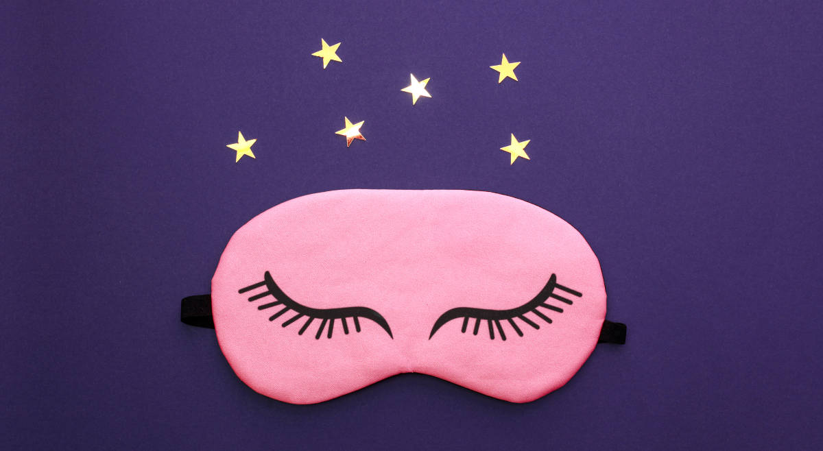 Pinke Schlafmaske mit draufgezeichneten geschlossenen Augen auf lila Hintergrund mit Sternen darüber.