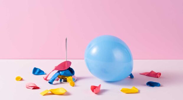 Ein blauer Luftballon vor rosa Hintergrund, daneben Reste bunter Luftballons, die von einer Nadel zerstochen wurden