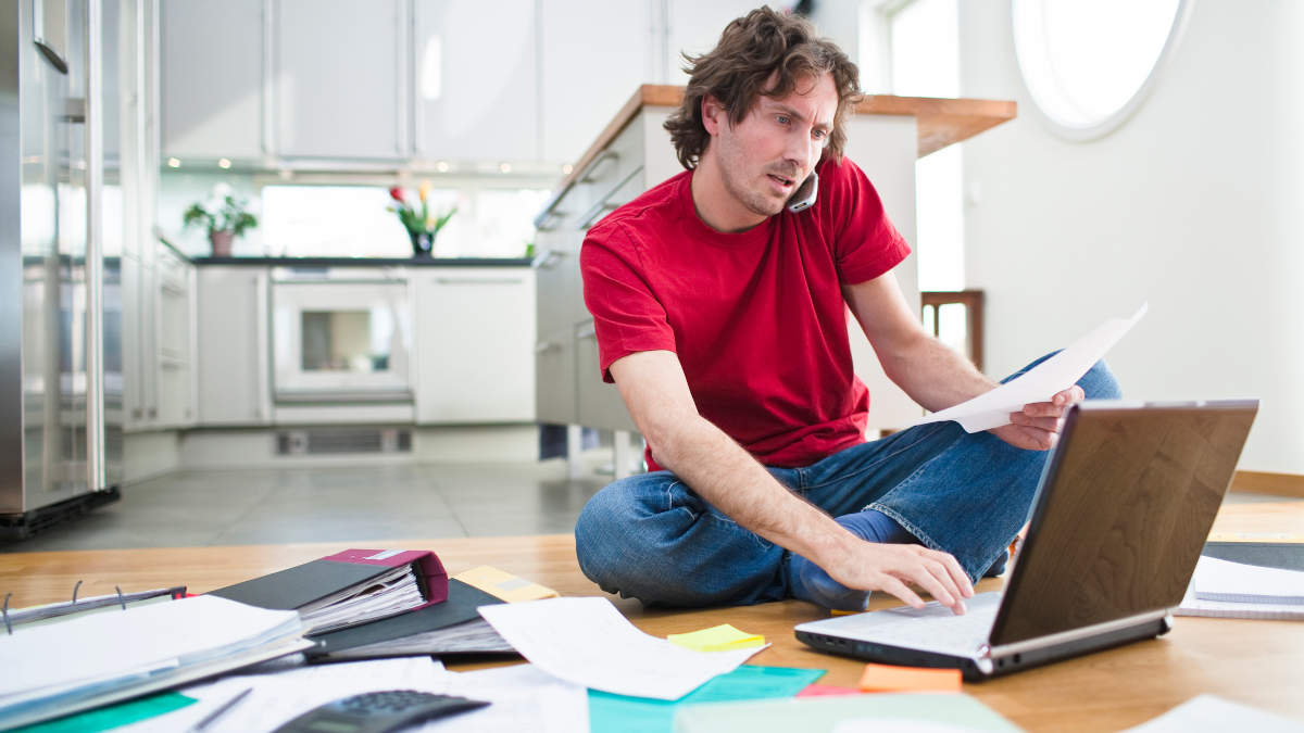 Mann im Homeoffice auf dem Boden mit Laptop in der Hand und Zettel-Chaos um ihn herum.