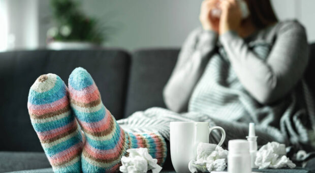 Frau sitzt auf Couch und putzt sich die Nase, die Ansicht ist verschwommen. Klar im Vordergrund sehen wir ihre Füße in Wollsocken, Medikamente, gebrauchte Taschentücher und einen Becher mit Tee.