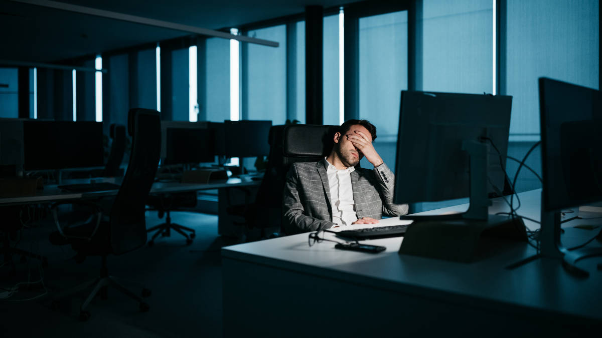 Businessmann sitzt vor Computer im Büro in der Dunkelheit und hält sich das Gesicht mit einer Hand zu. Rest des Büros ist dunkel und leer.