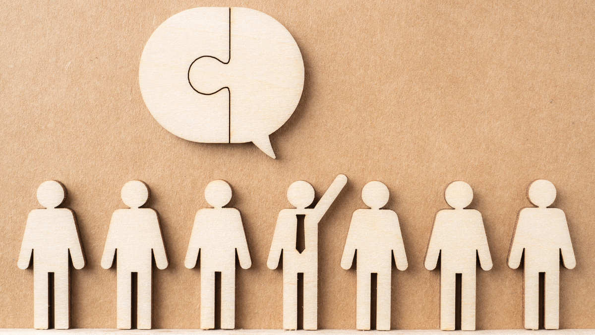 Mehrere Holzfiguren stehen nebeneinander, die Figur in der Mitte trägt Krawatte und macht eine Ansage.