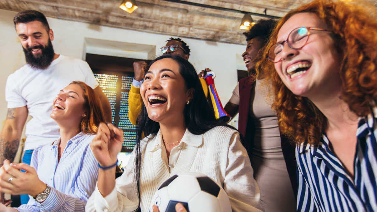 6 lachende Menschen in neutralem Raum. Frau im Vordergrund mit Fußball im Arm.