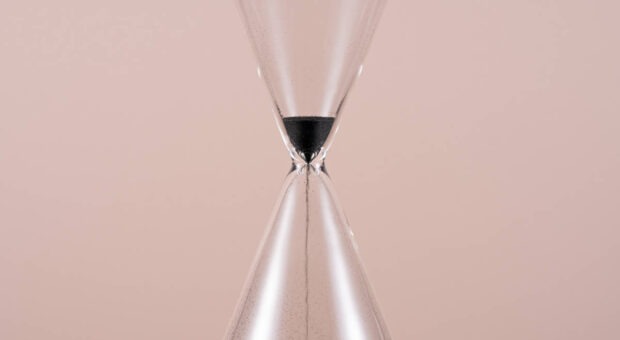 Mitte einer Sanduhr mit fast durchgelaufenem Sand auf rosa Hintergrund.