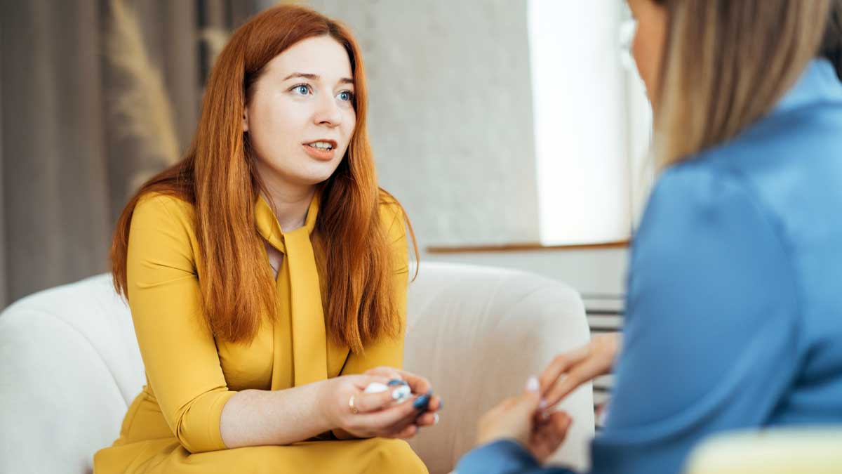 Eine Frau im gelben Kleid sitzt einer Frau in einer hellblauen Bluse gegenüber.