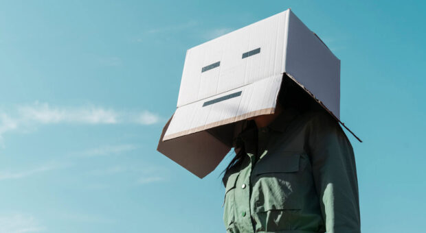 Eine Frau mit Pappkarton auf dem Kopf, auf den Karton ist ein unfreundlich schauender Smiley zu sehen