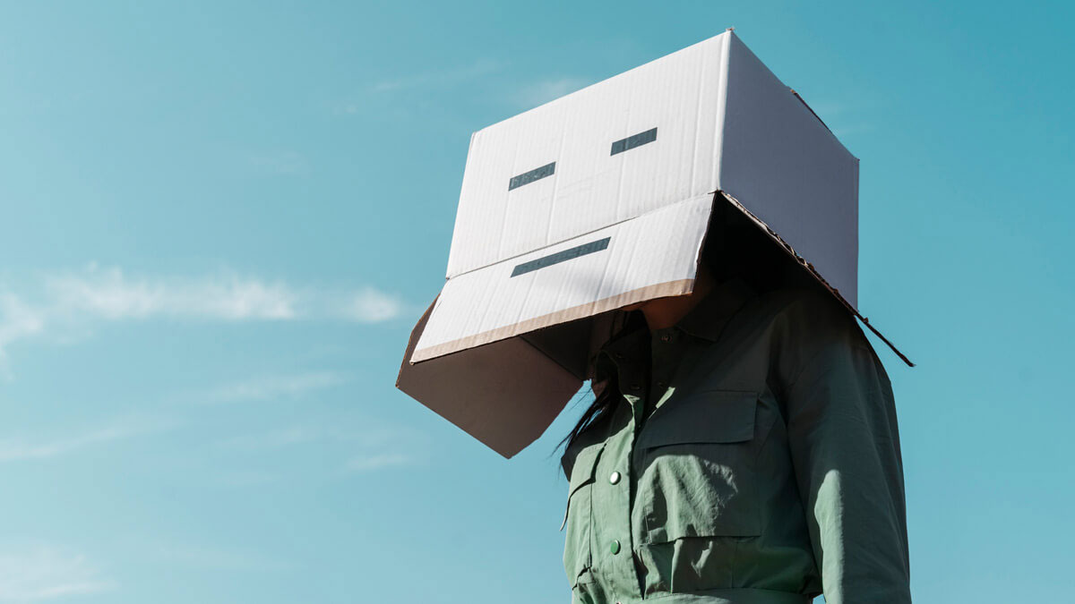 Eine Frau mit Pappkarton auf dem Kopf, auf den Karton ist ein unfreundlich schauender Smiley zu sehen