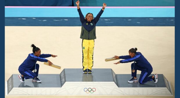 Das Bild zeigt die Siegerehrung der Bodenturnerinnen bei den Olympischen Spielen 2024. Siegerin ist Rebeca Andrade, Simone Biles (Silber) und die Drittplatzierte verneigen sich auf dem Siegerinnenpodest vor der Gold-Gewinnerin.