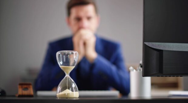 Eine Sanduhr steht auf eine Büro-Arbeitstisch, im Hintergrund ist etwas verschwommen ein Mann zu sehen