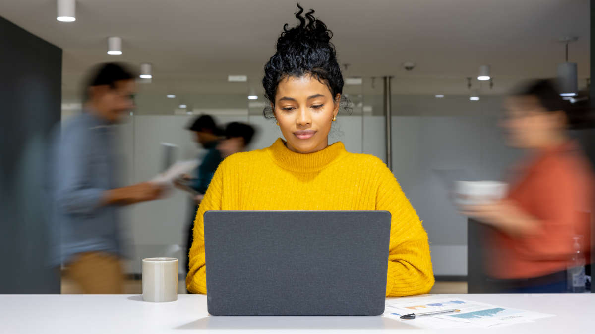Eine Frau am Laptop in einer Büroumgebung, die im Flow zu sein scheint, während um sie herum verschwommen dargestellte Kolleginnen und Kollegen hin- und herlaufen.