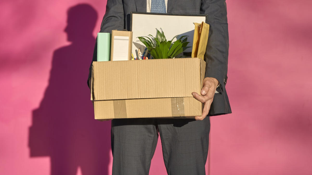 Person, die einen Karton mit Büroartikeln in den Händen hält, vor einer pinken Wand.