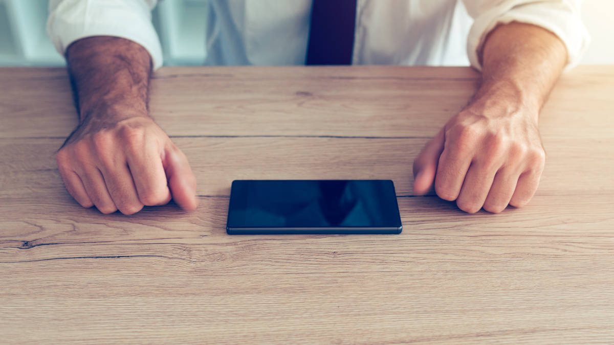 Mann im Anzug der an einem Tisch sitzt. Sein Smartphone liegt in der Mitte des Tisches und er hat die Hände, die zu Fäusten geballt sind, rechts und link von Smartphone liegen.