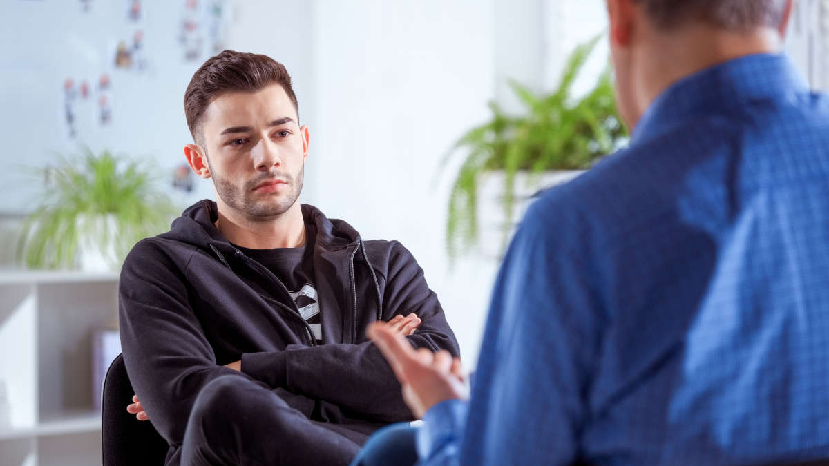 Ein junger, deprimiert guckender Mann mit verschränkten Armen hört einem älteren Mann zu.