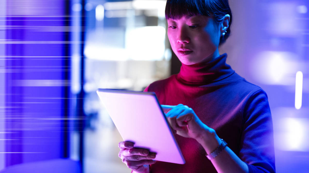 Frau steht in einem Büro und hält ein Tablet in der einen Hand und tippt mit der anderen Hand. Die Farbwahl des gesamten Bildes sind Blau- und Rottöne