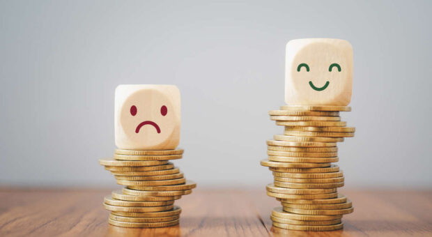 Ein Würfel mit traurigem Smiley auf einem niedrigen Stapel Münzen und ein Würfel mit fröhlichem Smiley auf einem höheren Stapel Münzen.