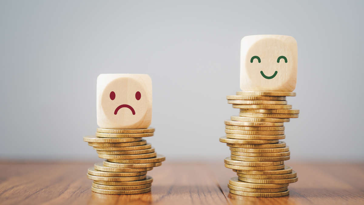 Ein Würfel mit traurigem Smiley auf einem niedrigen Stapel Münzen und ein Würfel mit fröhlichem Smiley auf einem höheren Stapel Münzen.