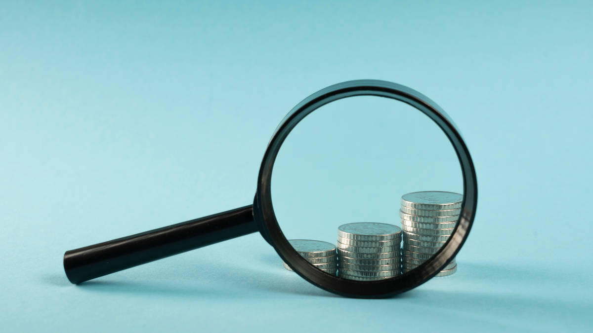 Drei verschieden hohe Türme aus Münzen hinter einer Lupe.