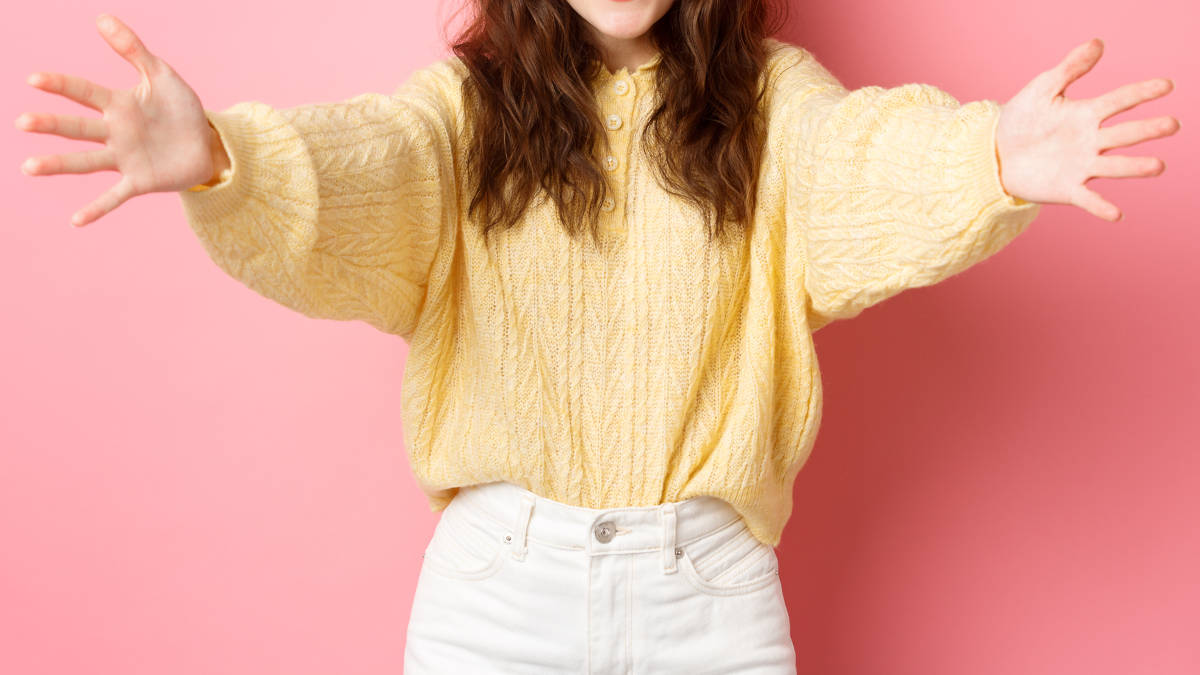 Frau in gelbem Pulli und weißer Hose, die die Arme zu einer Umarmung ausbreitet