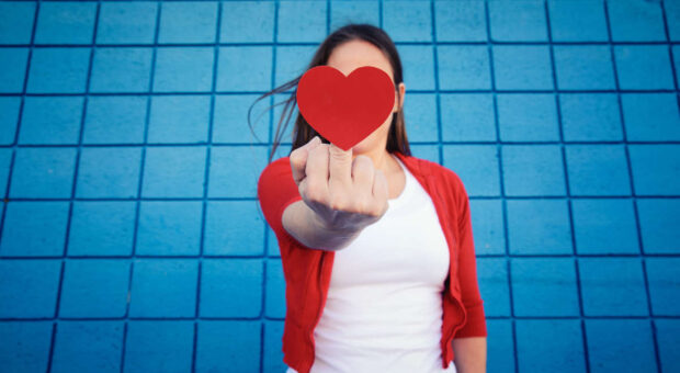 Eine Person streckt dem Betrachter ihren Mittelfinger entgegen, an dem aber ein rotes Herz befestigt ist