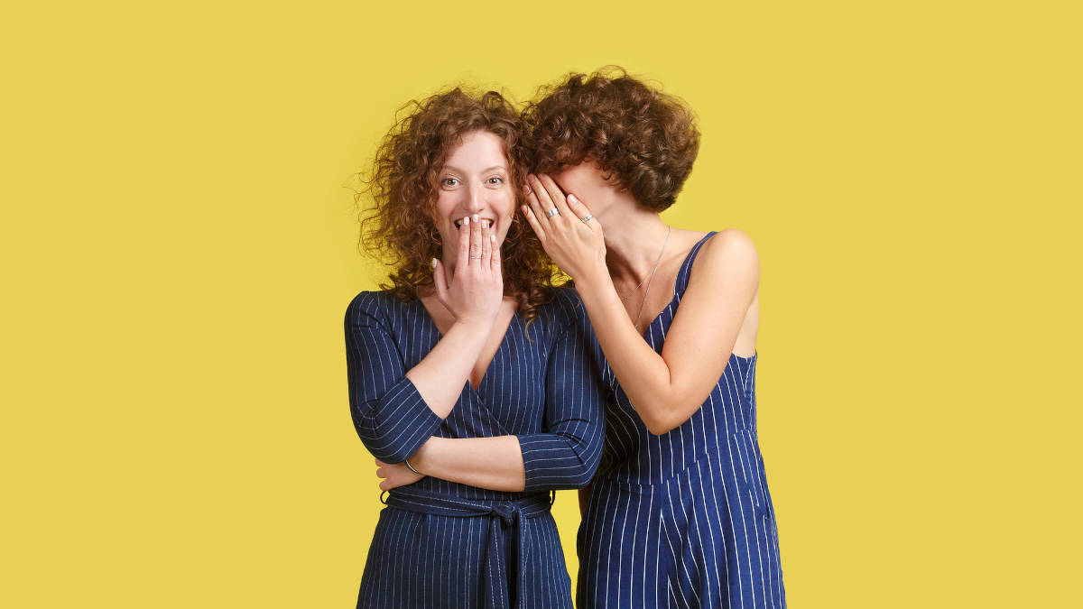 Zwei Frauen vor einem gelben Hintergrund. Die Frau rechts flüstert der Frau links etwas ins Ohr - diese lacht.