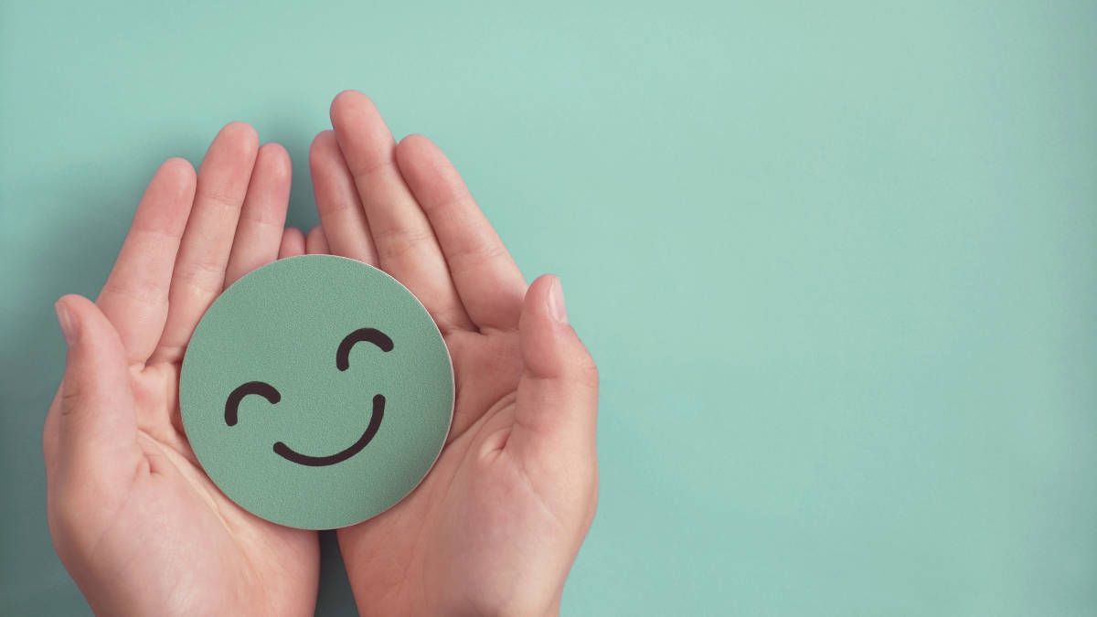 Zwei Hände formen eine Schale und halten darin einen grünen, lächelnden Smiley aus Papier.