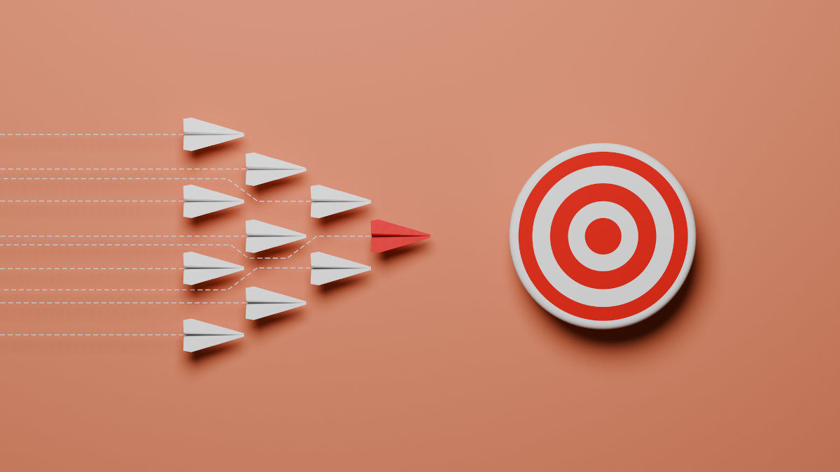 Eine Formation an weißen Papierfliegern fliegt in Richtung einer rot-weißen Zielscheibe. Der Papierflieger an der Spitze der Formation ist rot.