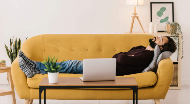 Mann liegt auf Couch mit geschlossenen Augen. Sein Laptop steht vor ihm auf dem Couchtisch