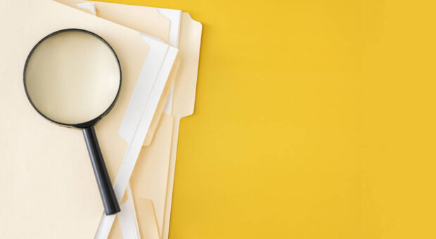 Eine Lupe liegt auf einem Stapel Unterlagen vor knallig gelbem Hintergrund
