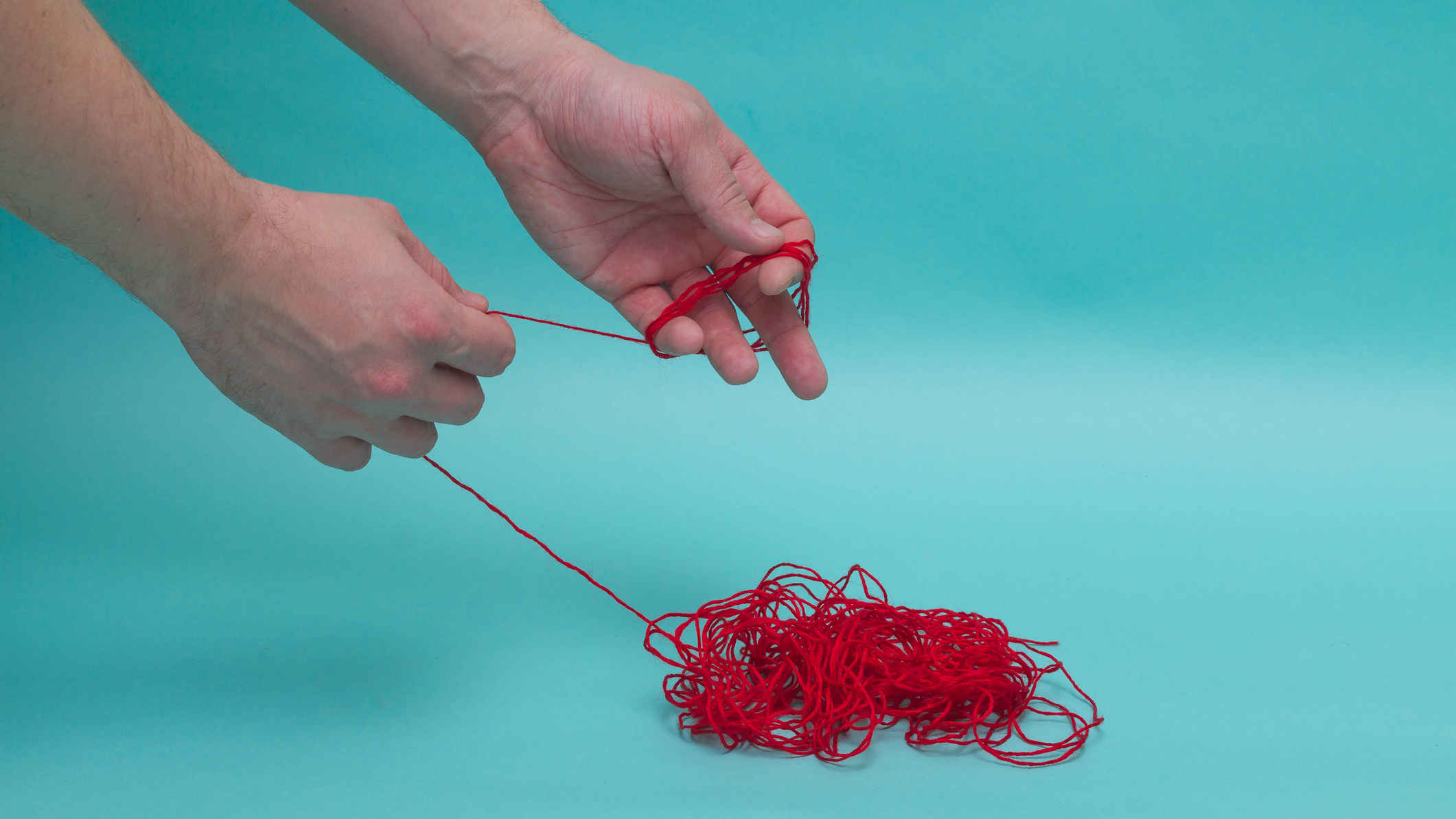 Ein Mensch entzerrt ein rotes Knäuel Wolle vor türkisem Hintergrund. Zu sehen sind vom Menschen nur die Hände.