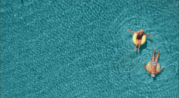 Vogelperspektive: Zwei ältere Menschen liegen in großen Schwimmreifen in klarem Wasser