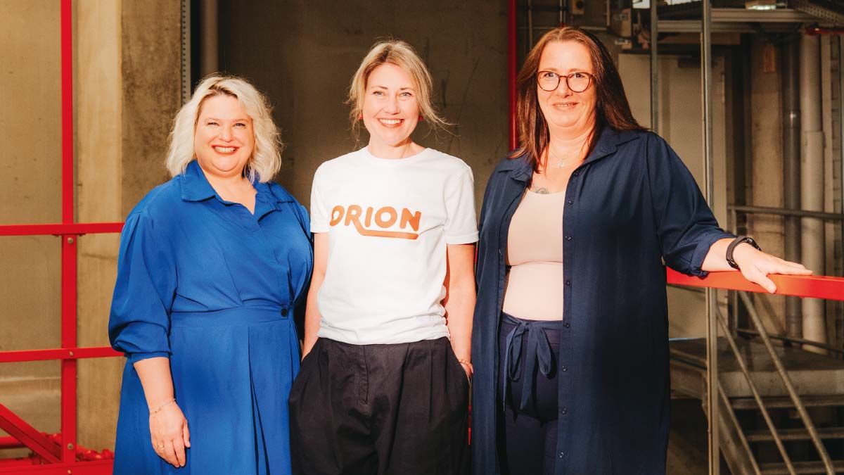 Drei Frauen stehen lächelnd nebeneinander. Die mittlere Frau trägt ein weißes Shirt mit der Aufschrift "Orion".