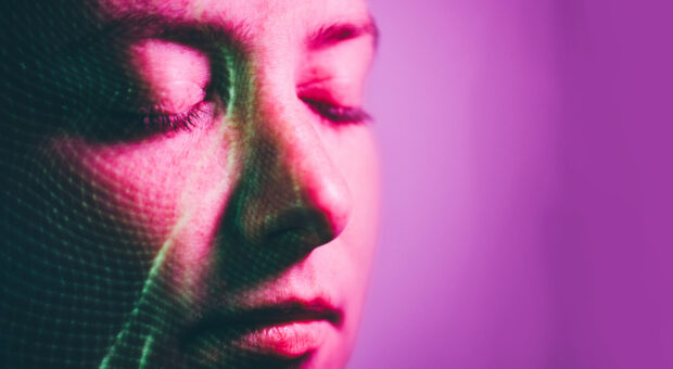 Menschliches Gesicht mit geschlossenen Augen in Nahaufnahme. Auf dem Gesicht sind grünliche Lichtspuren zu sehen. Der Hintergrund des Bildes ist lila.