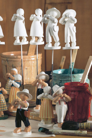 Verschiedene Holzfiguren, von denen einige bemalt sind, andere noch nicht. Im Hintergrund stehen offene Farbtöpfe.